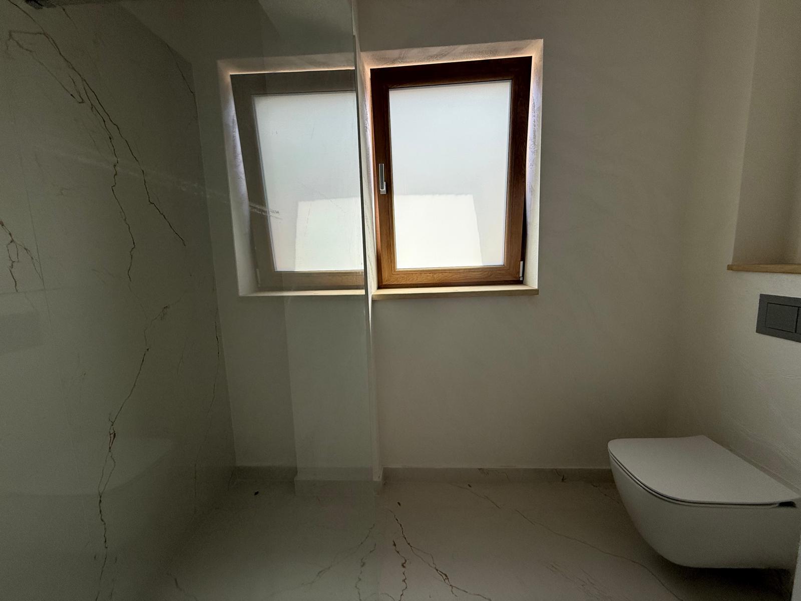 Baie modernă cu faianță tip marmură, toaletă suspendată și fereastră mate din lemn