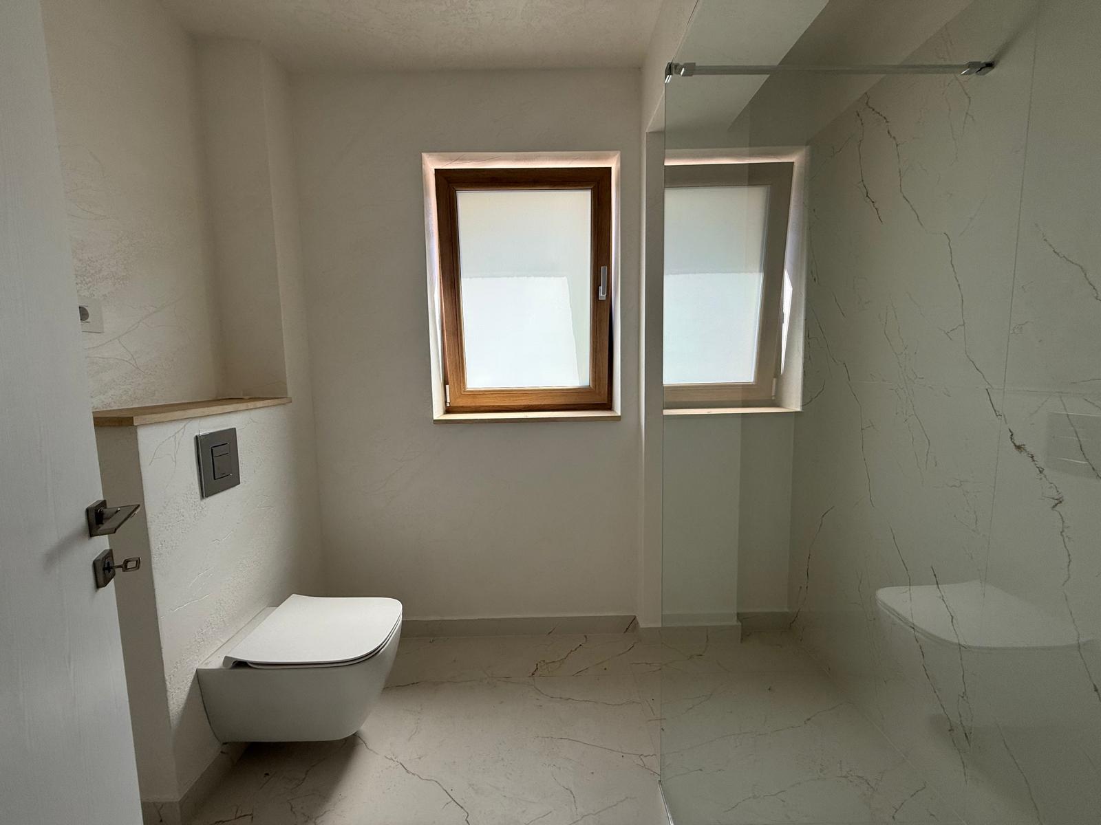 Baie modernă cu duș walk-in din sticlă, toaletă suspendată și fereastră mascată
