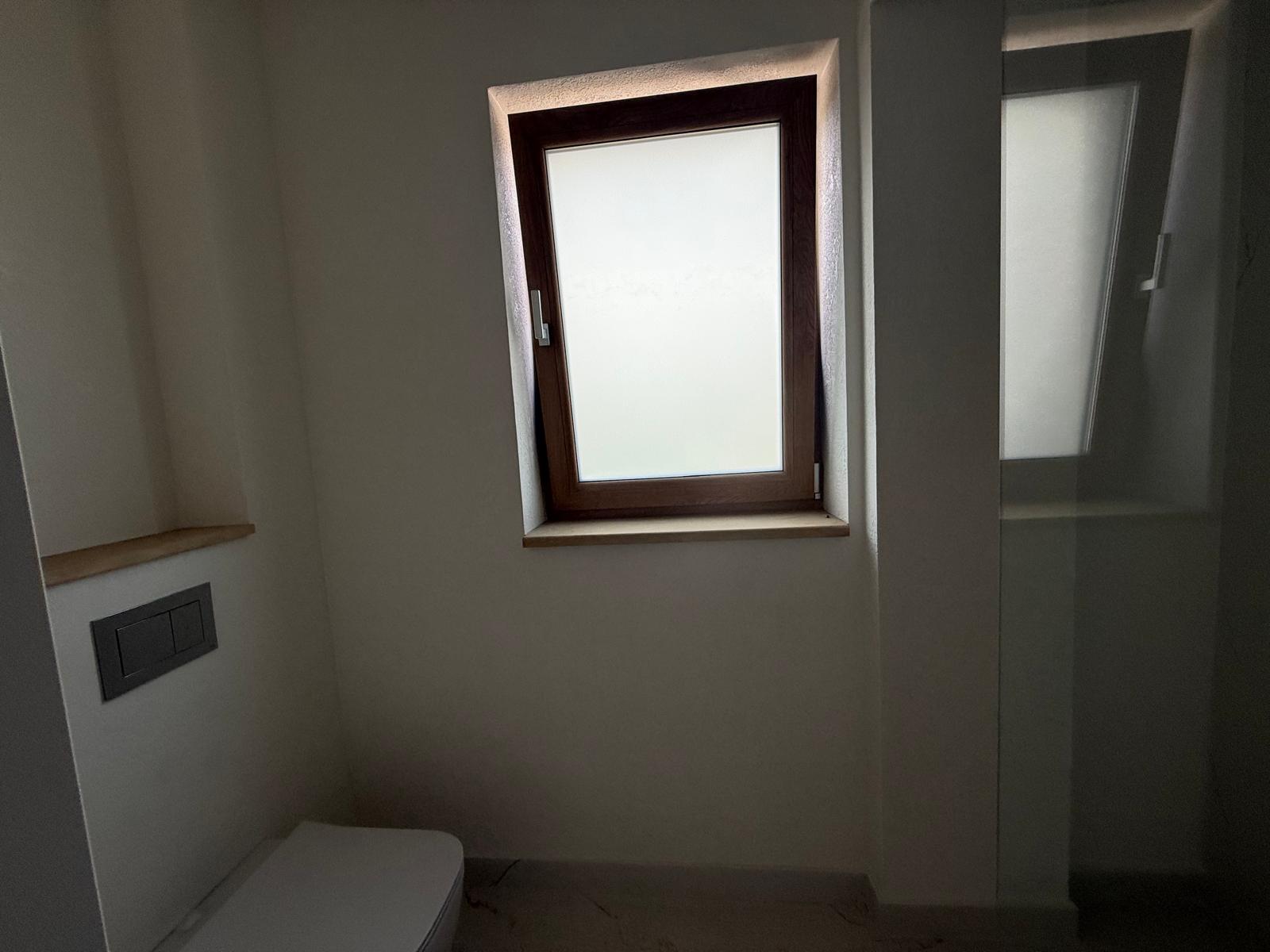 Baie modernă cu toaletă suspendată, cabină de duș din sticlă și fereastră mate cu rama din lemn