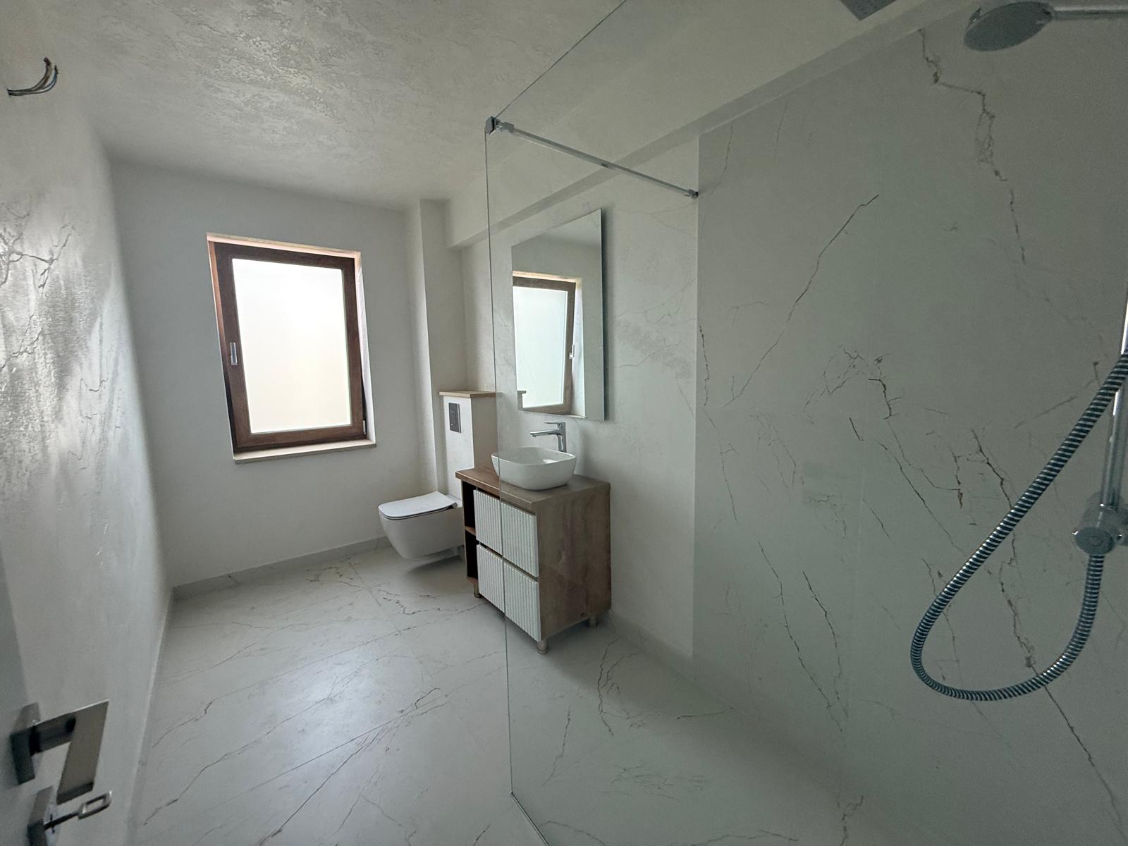 Baie modernă cu duș walk-in din sticlă, marmură, toaletă suspendată și dulap din lemn
